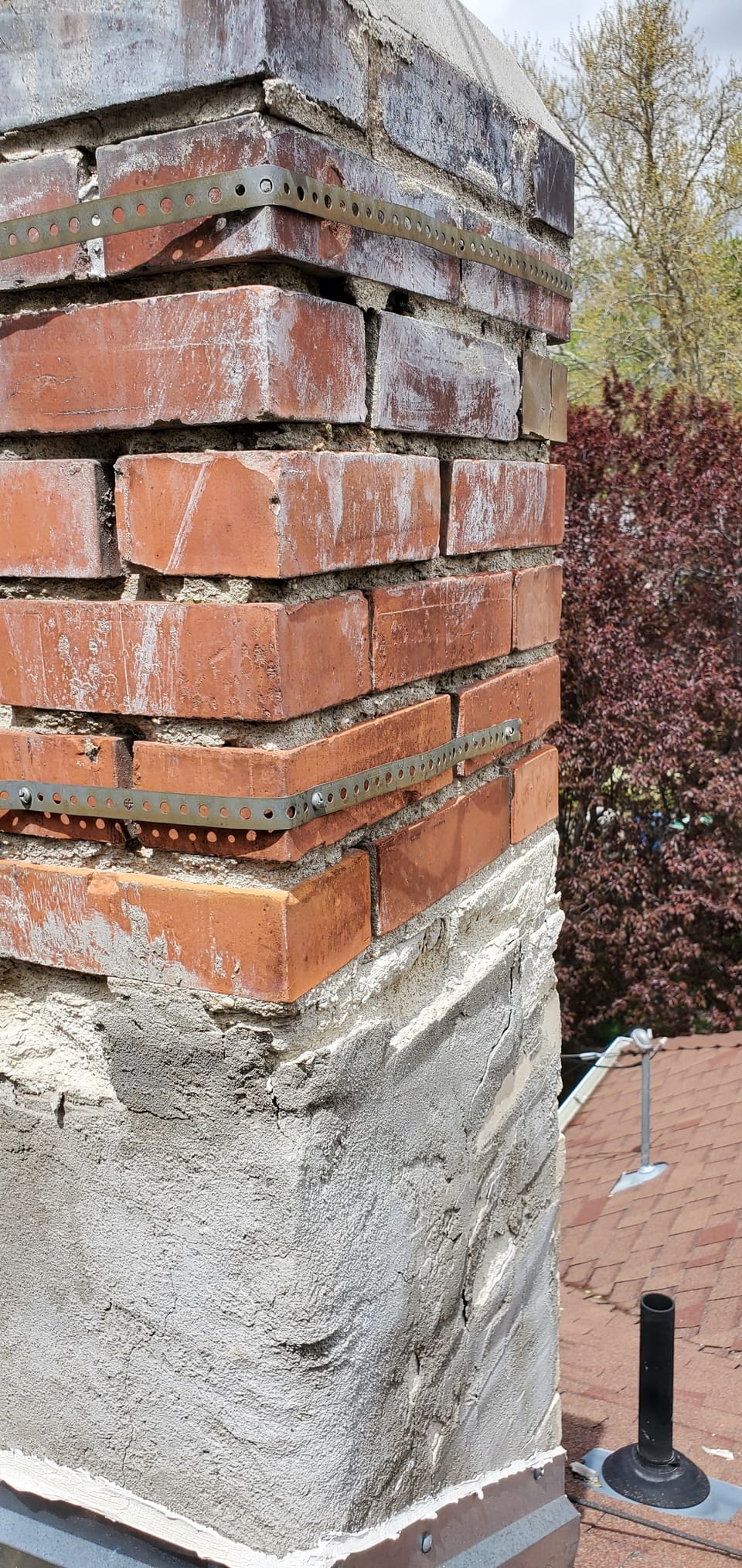 Failed stucco coat cracking and falling away below brick with washed-out mortar joints and metal strapping