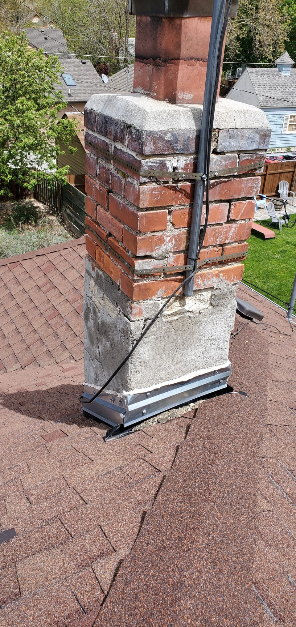 Original brick chimney with metal strapping wrapped around it and a cracked cap above a red shingle roof