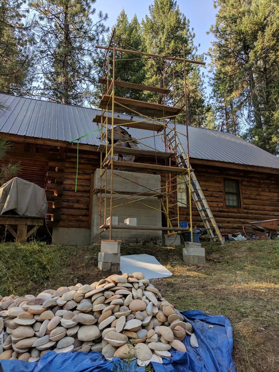Scaffold set against the log cabin chimney chase with manufactured river rock piled on a tarp below