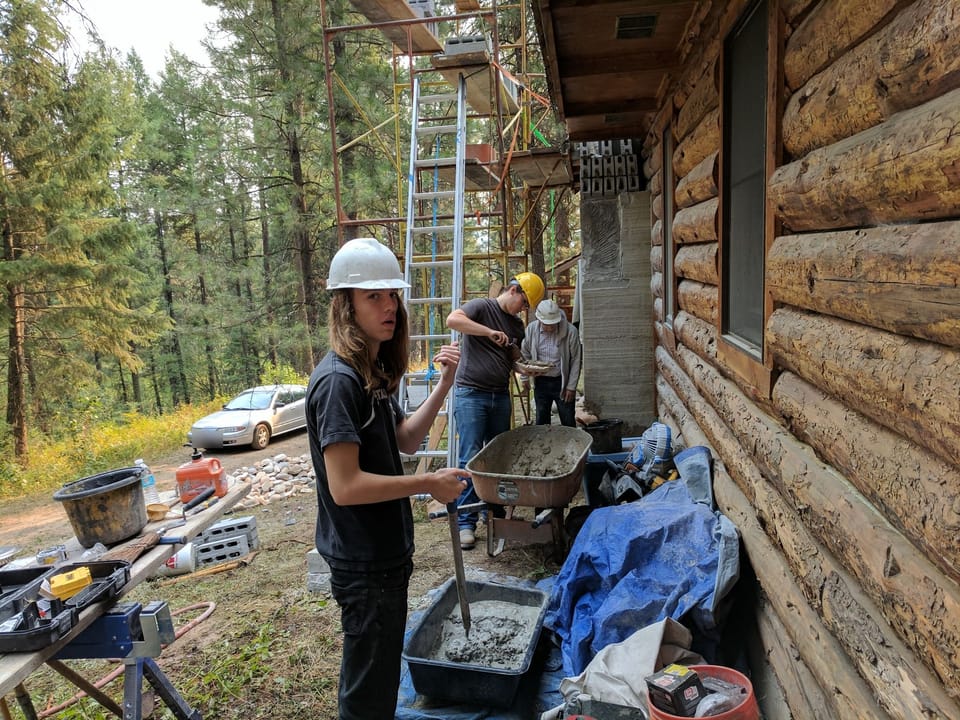 Apprentice mixing mortar in a tub while two masons work the chimney chase on the log cabin