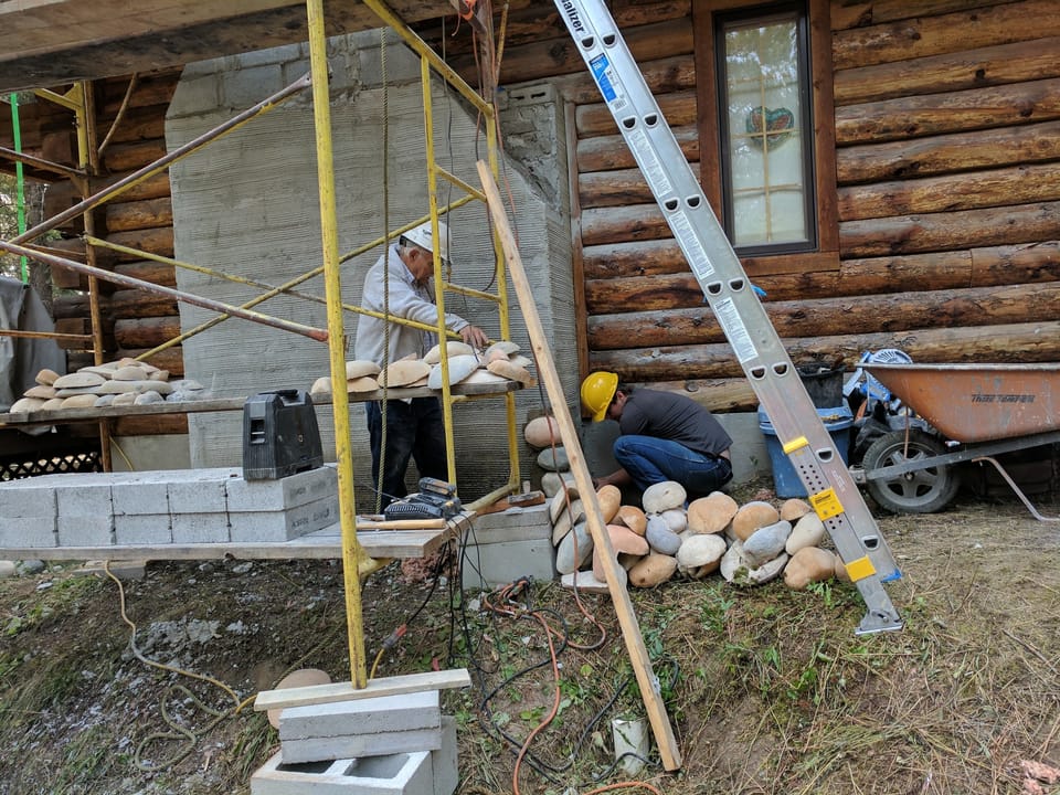 Older mason sorting manufactured river rock on a plank while an apprentice sets stone at the chimney base