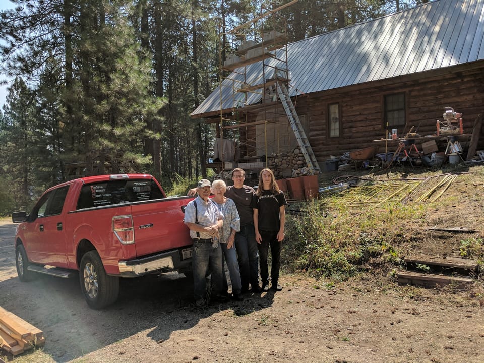 Four family members beside the red Fackrell Masonry pickup at the log cabin, scaffold on the chimney behind