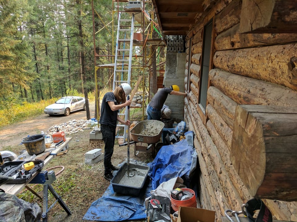 Apprentice mixing mortar with a paddle mixer next to the scratch-coated chimney chase on the log wall