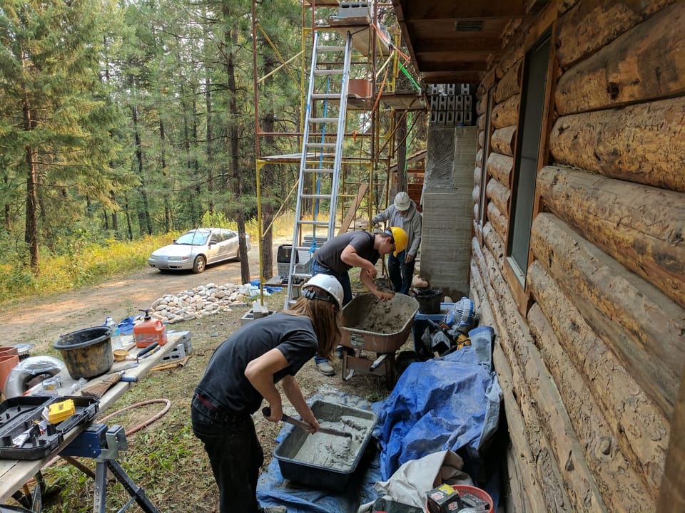 Two apprentices working mortar into a wheelbarrow and tub beside the log cabin chimney chase