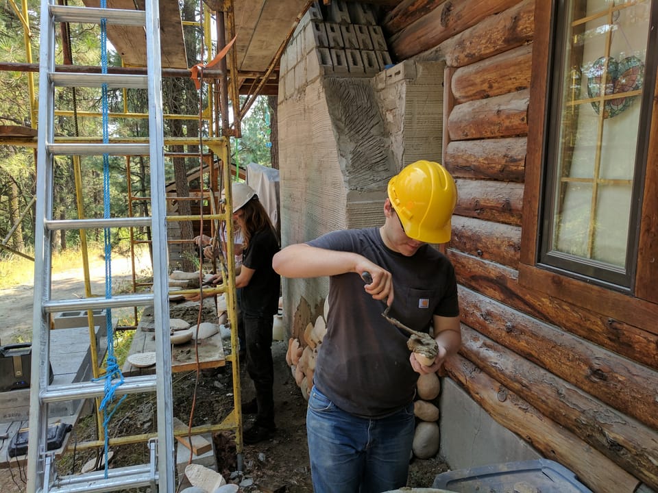 Apprentice buttering the back of a river rock with a trowel beside the scratch-coated chimney chase