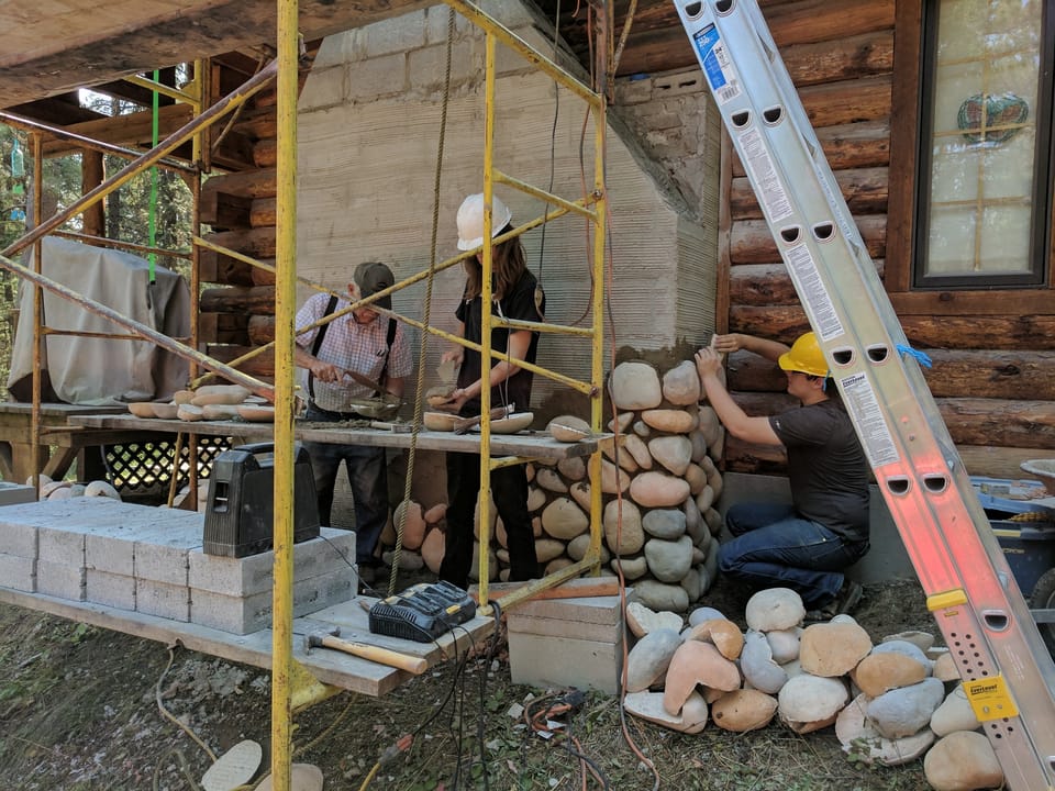 Three masons working the chimney chase as river rock veneer reaches waist height on the log cabin