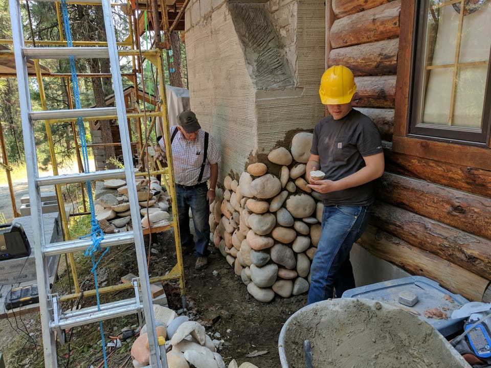 manufactured river rock laid chest high on the log cabin chimney, apprentice holding the next stone