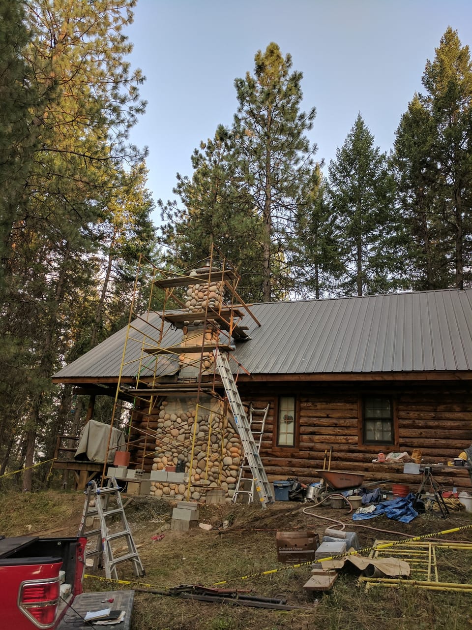 manufactured river rock chimney built past the metal roof to the cap with scaffold still standing against the cabin