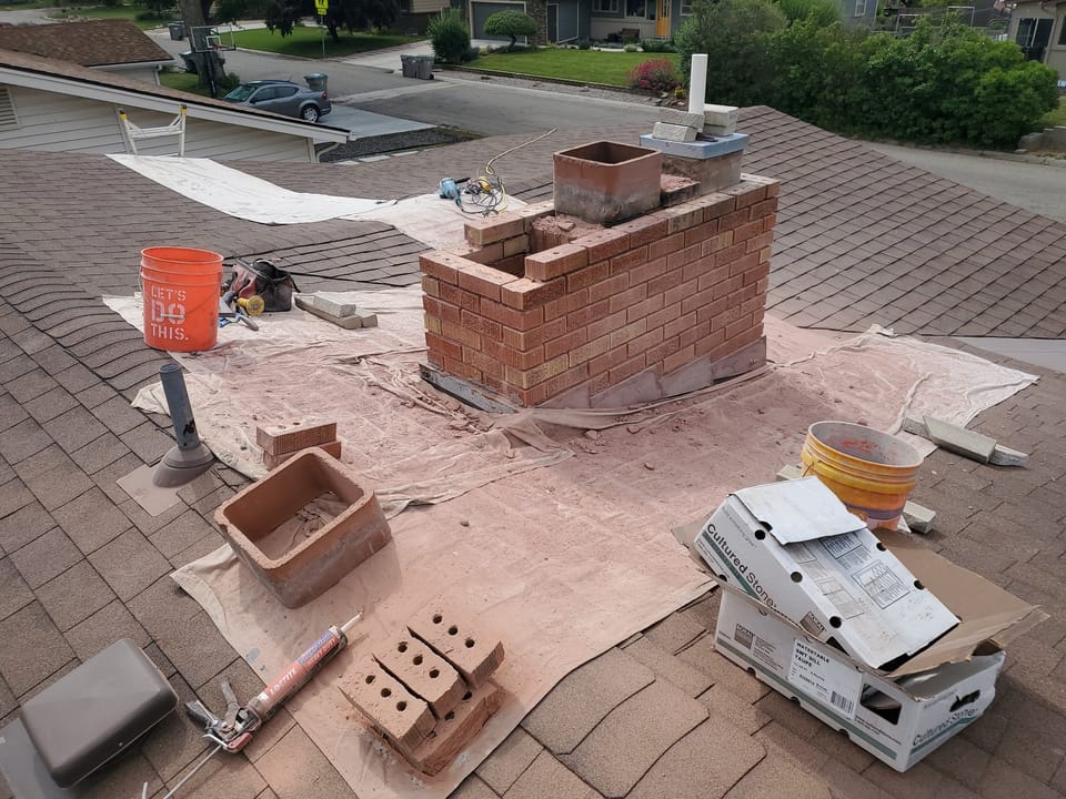 Chimney rebuild in progress on a shingle roof with drop cloths, fresh brick courses and flue tile