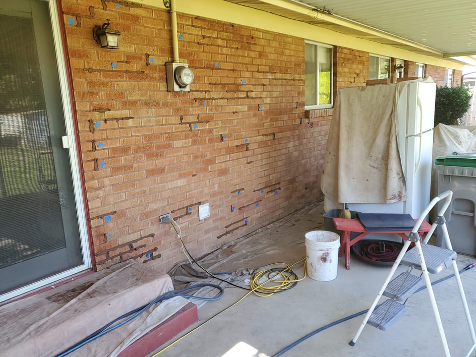 Brick wall under a covered patio with mortar joints ground out and repointed, blue tape marking work