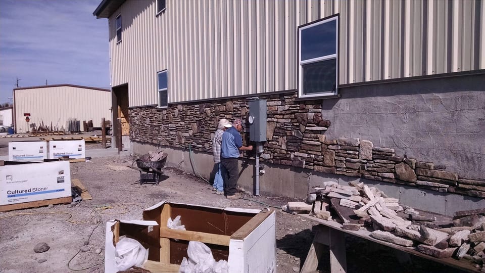 Two people at the new stone veneer wainscot beside the electrical meter, cut stone stacked on a table