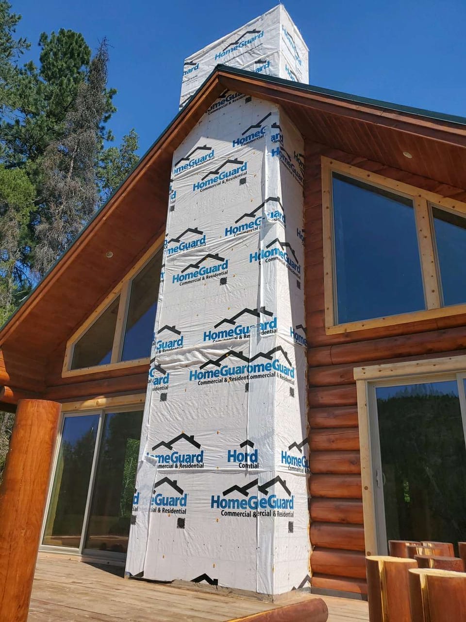 Framed chimney chase wrapped in white housewrap rising past the roofline of a log home, ready for stone