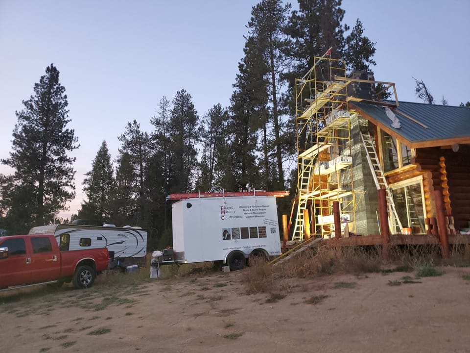 Scaffold and a Fackrell Masonry and Construction trailer beside a log home at dusk, stone veneer partly installed