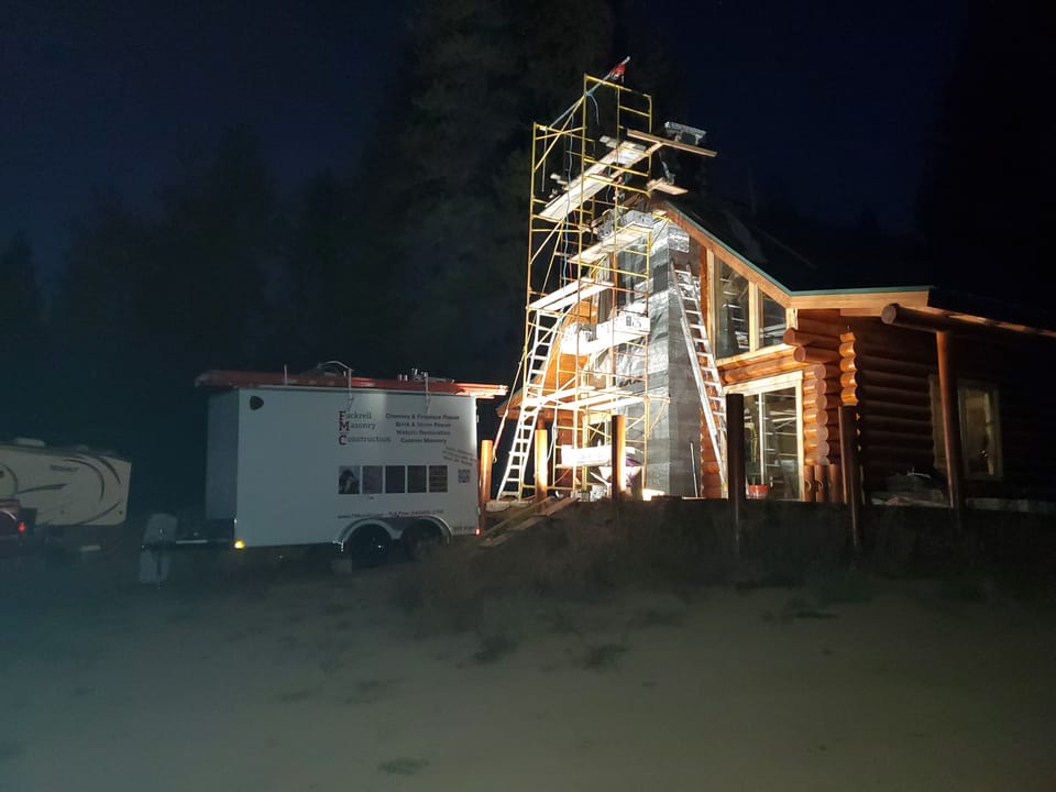 Floodlit scaffold against the chimney of a log home after dark, work continuing at night