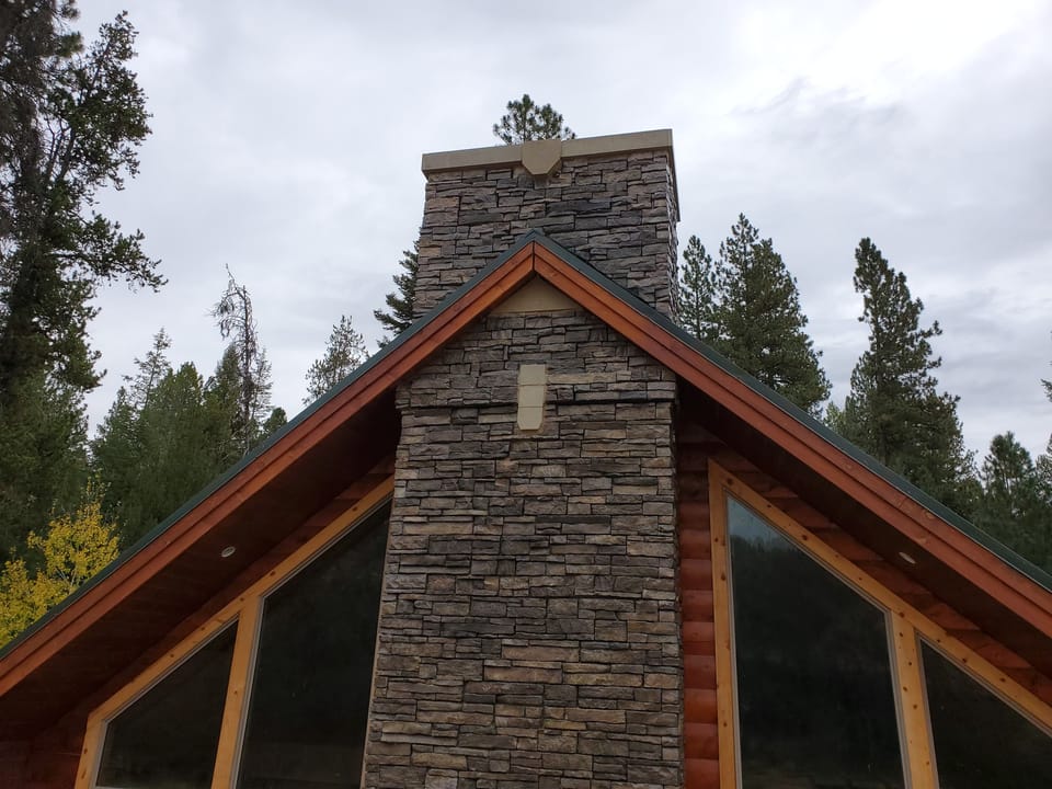 Finished stone veneer chimney with a cast stone cap, seen from below against pines and sky