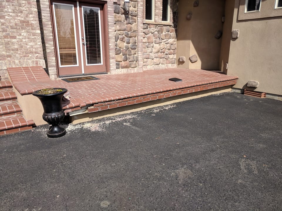 Old brick patio with crumbling edge bricks and a high step down to the asphalt driveway