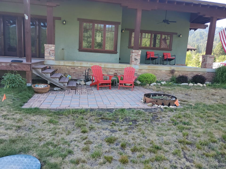 Small original brick patio with red Adirondack chairs and a fire ring behind a green craftsman home