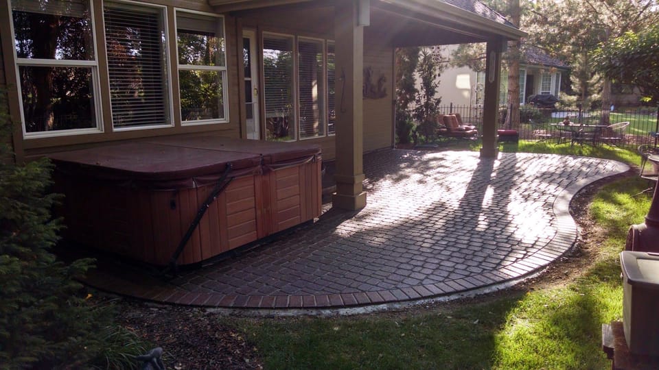 Sealed paver patio with a brick soldier-course border and deep wet-look color beside a hot tub