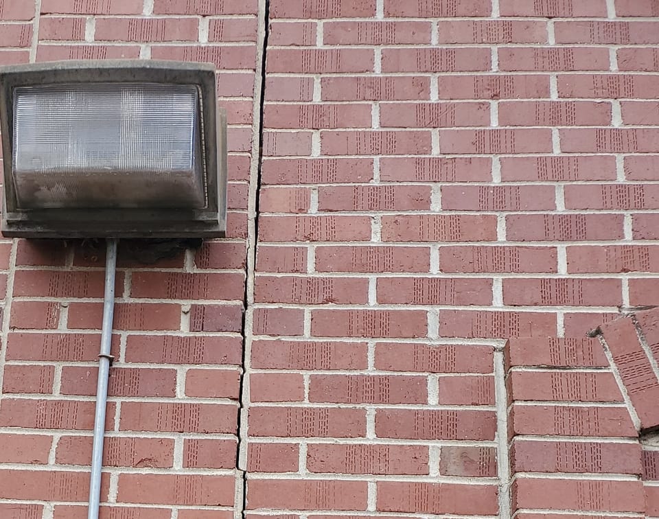 Open vertical crack running through the mortar joints of a red brick wall beside a wall-pack light