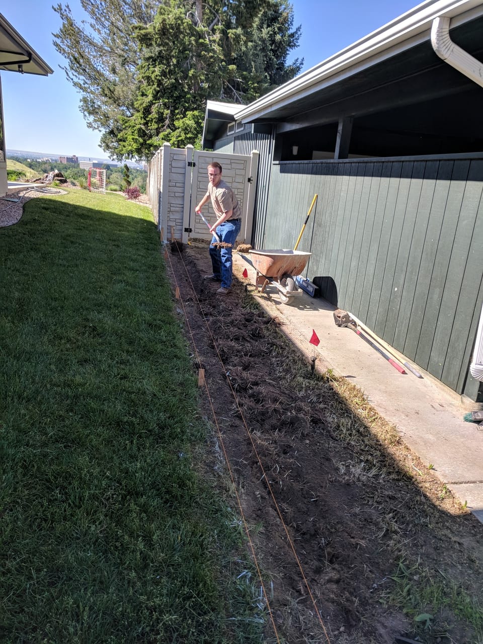 Hand-digging a footing trench with a shovel along a fence line, string lines and marking flags set