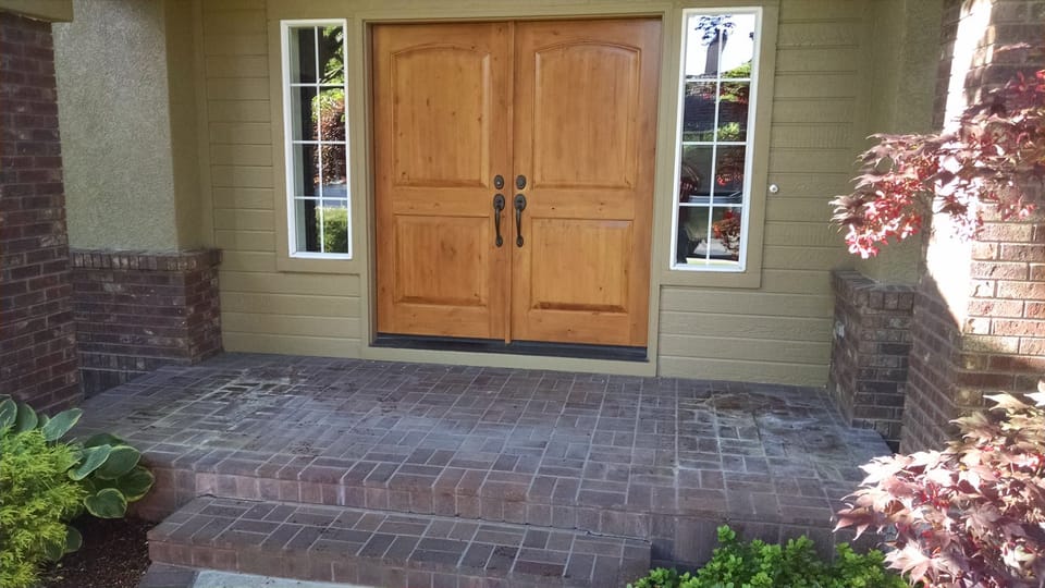 Brick front porch and step covered in dark stains and pale blotches before cleaning, wood entry doors behind