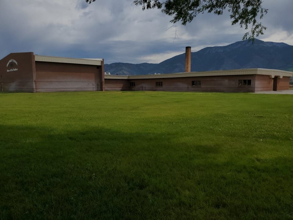 Swan Valley School brick building across the lawn under storm clouds, lower brick band darkened by sprinkler staining