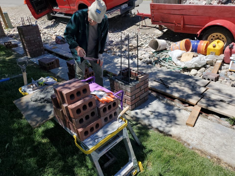 Older mason in a flannel shirt trowelling mortar on a rebuilt brick fence post in Arco, Idaho